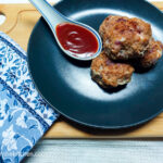 On a white background, a wooden cutting board, in the left hand lower corner a blue patterned cloth napkin. In the middle of the photo, a black, matte plate with a stash of meatballs, and a porcelain spoon with barbecue sauce.