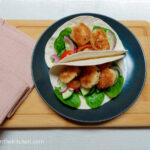 On a white textured background, a wooden cutting board, with a pink cloth napkin on the left side and a black matte plate in the middle. The plate is filled with two soft shell tacos, each have a few spinach leaves on the bottom, topped with red onions, tomatoes and cucumbers, and on the top, there are crispy prawns.