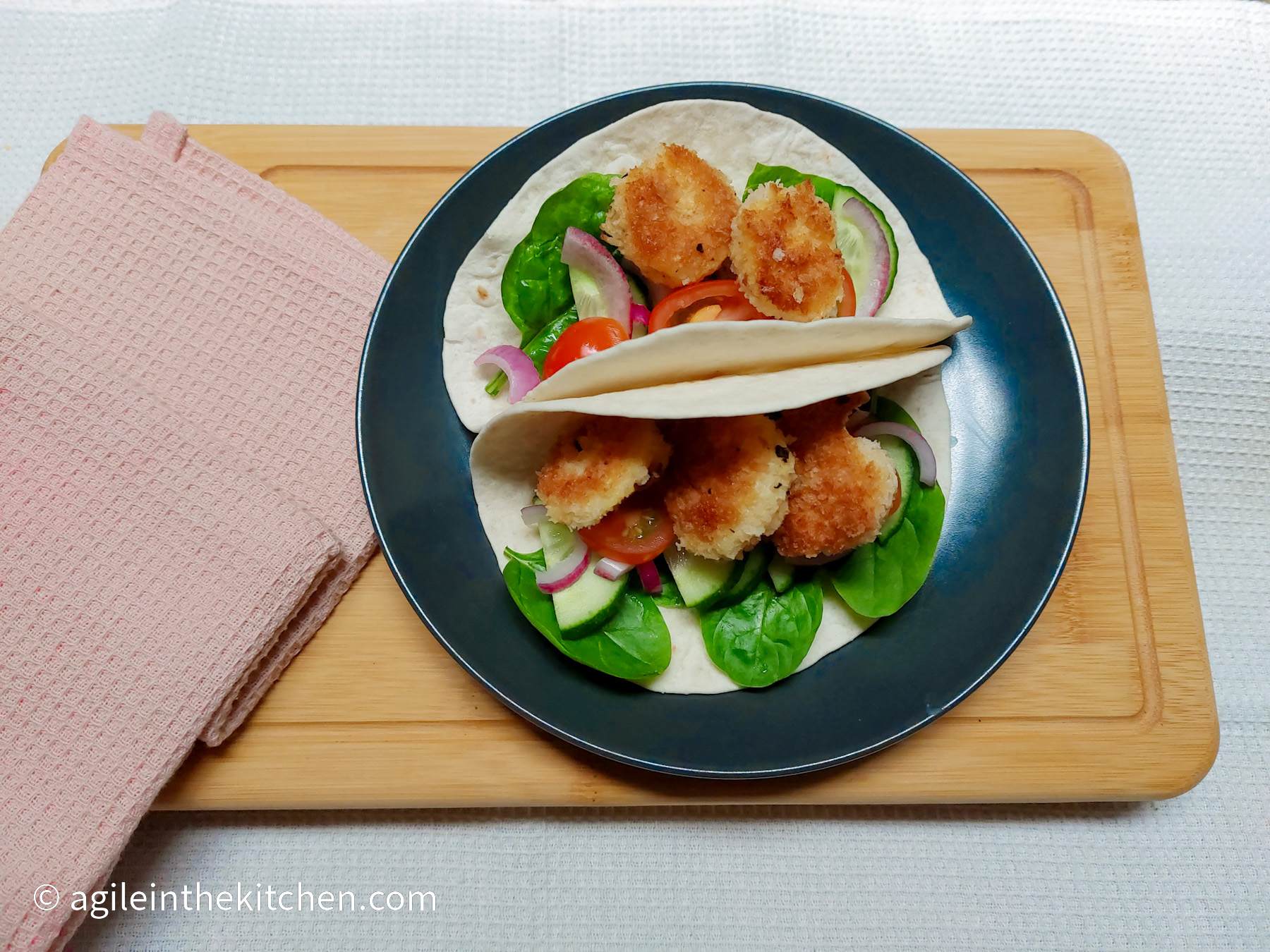On a white textured background, a wooden cutting board, with a pink cloth napkin on the left side and a black matte plate in the middle. The plate is filled with two soft shell tacos, each have a few spinach leaves on the bottom, topped with red onions, tomatoes and cucumbers, and on the top, there are crispy prawns.