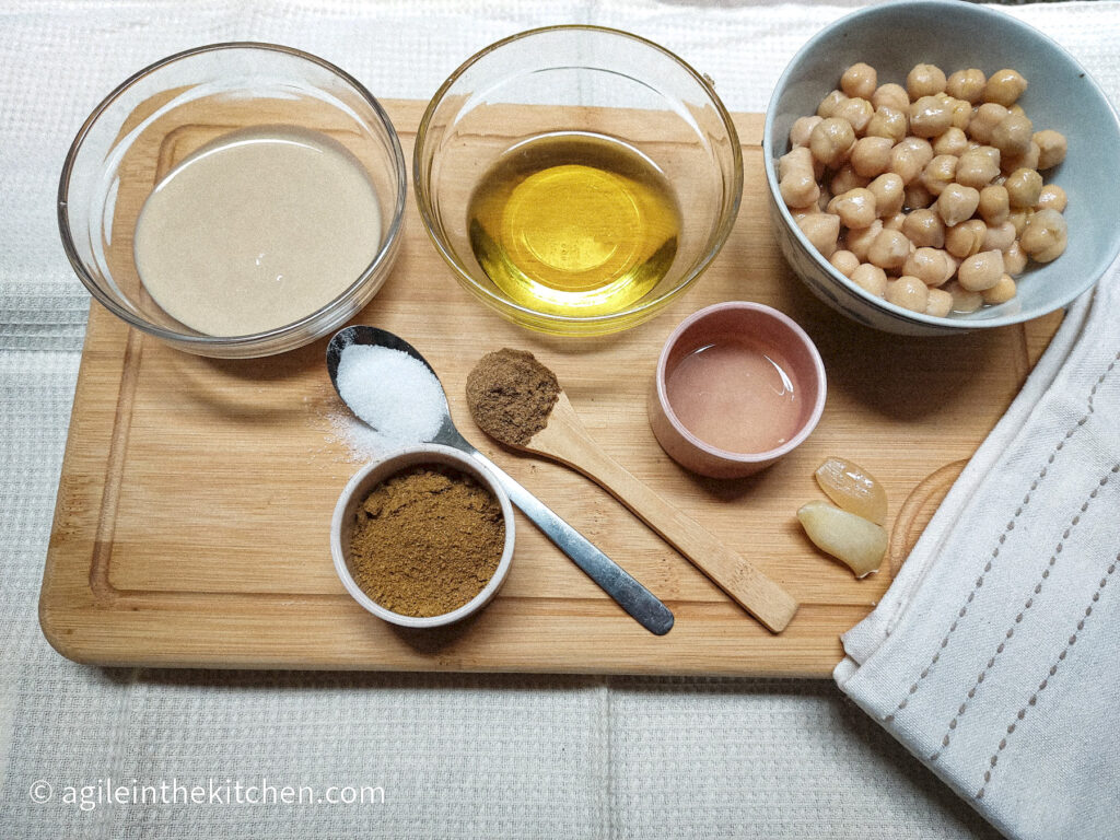 Laid out the ingredients to make falafelish dressing. Clockwise from upper left corner: a bowl of tahini, a bowl of olive oil, a bowl of cooked chickpeas, two cloves of garlic, a small, pink bowl of faba water, a wooden teaspoon of cumin, a teaspoon of salt and a small bowl of falafel spices.