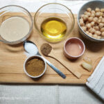 Laid out the ingredients to make falafelish dressing. Clockwise from upper left corner: a bowl of tahini, a bowl of olive oil, a bowl of cooked chickpeas, two cloves of garlic, a small, pink bowl of faba water, a wooden teaspoon of cumin, a teaspoon of salt and a small bowl of falafel spices.