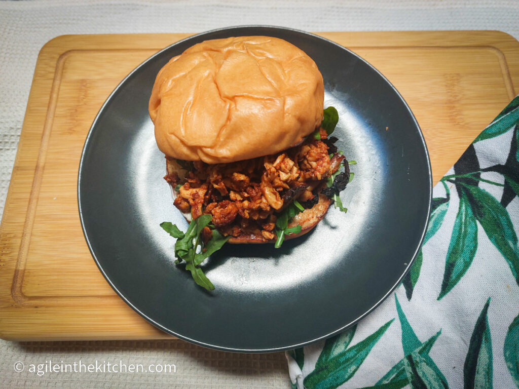 On a wooden cutting board, a black, matte plate with a hamburger bun filled with shredded barbecue tofu and a few lettuce leaves. In the right hand lower corner, a cloth napkin with a leaf print.