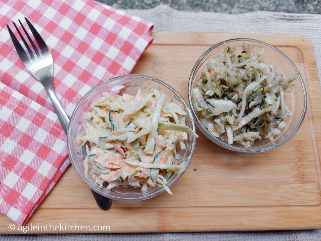 On a wooden cutting board, in the upper left hand corner a red gingham cloth napkin, in the middle a glass bowl with spicy coleslaw and one glass bowl with a white cabbage salad, with a fork to the left of the bowls