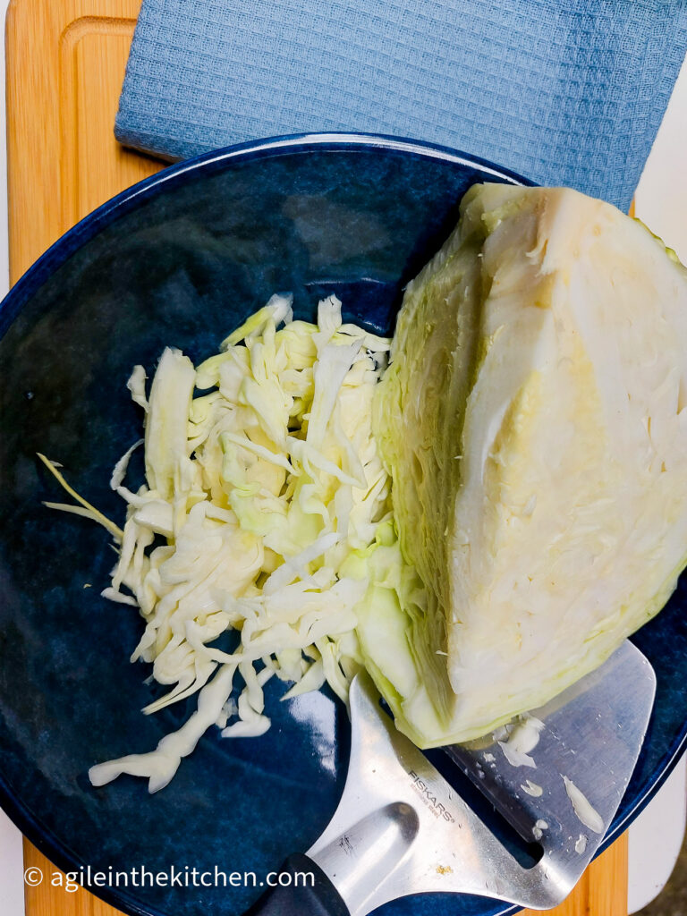 On a wooden cutting board, a blue, textured cloth napkin, a dark blue bowl with a white cabbage head and a cheese slicer, where some of the cabbage has already been sliced.