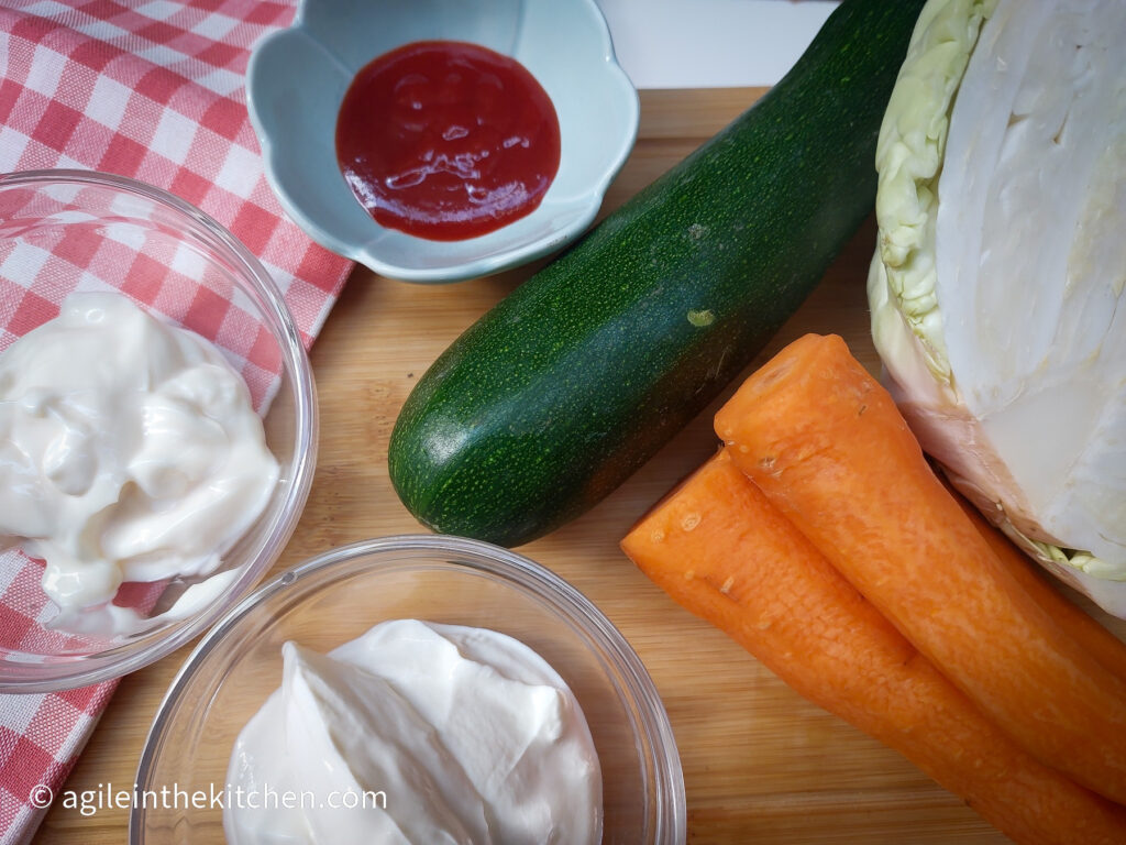 On a wooden cutting board with a red, gingham cloth in the upper right hand corner, ingredients to make spicy coleslaw is laid out, clockwise from top left, a flowershaped bowl with sriracha, a zucchini, half a head of white cabbage, two carrots, a glass bowl of Greek yogurt, a glass bowl of mayonnaise.
