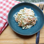 On a wooden cutting board, in the upper left hand corner a red gingham cloth napkin, in the middle a blue plate with spicy coleslaw, and a fork to the right of the plate