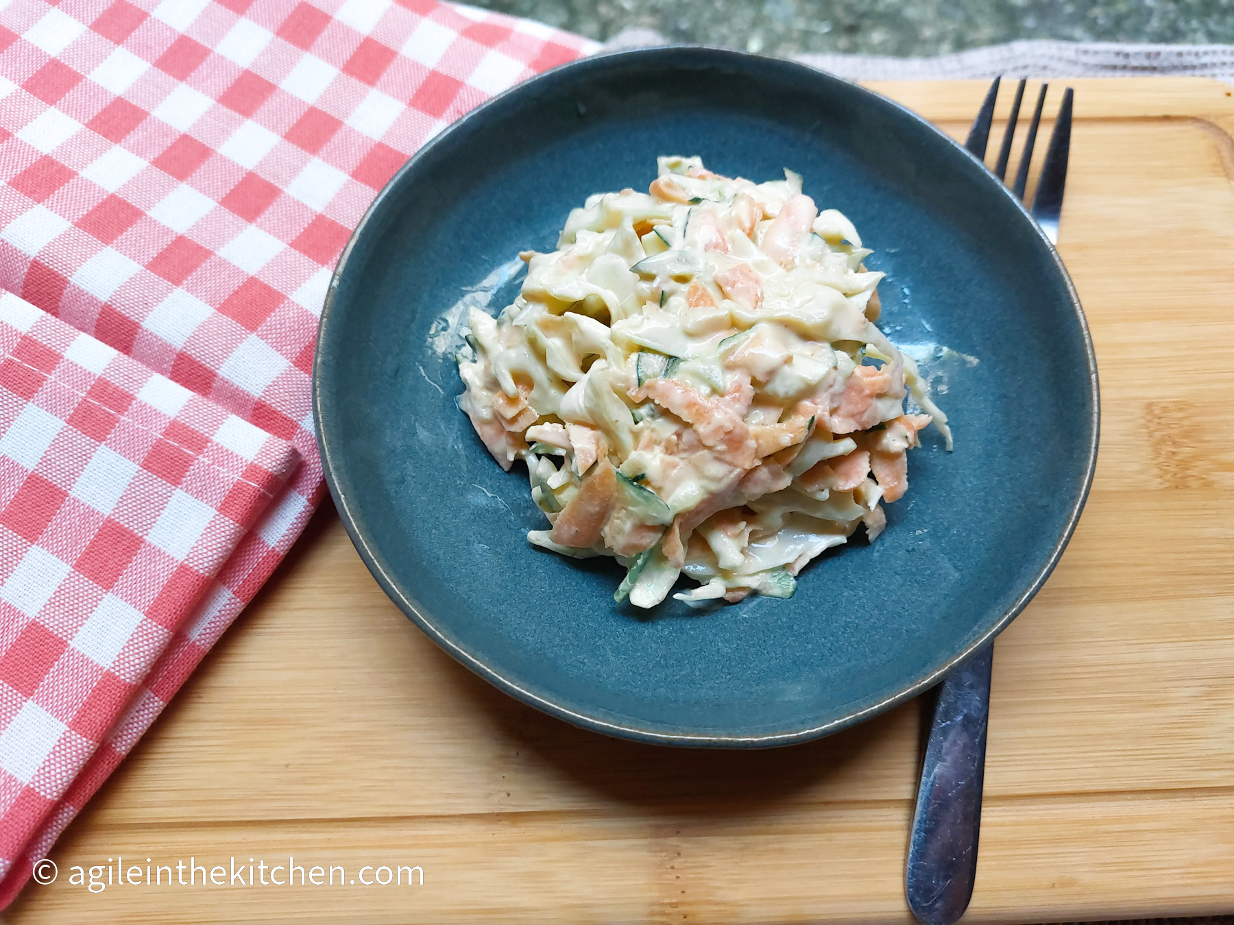On a wooden cutting board, in the upper left hand corner a red gingham cloth napkin, in the middle a blue plate with spicy coleslaw, and a fork to the right of the plate
