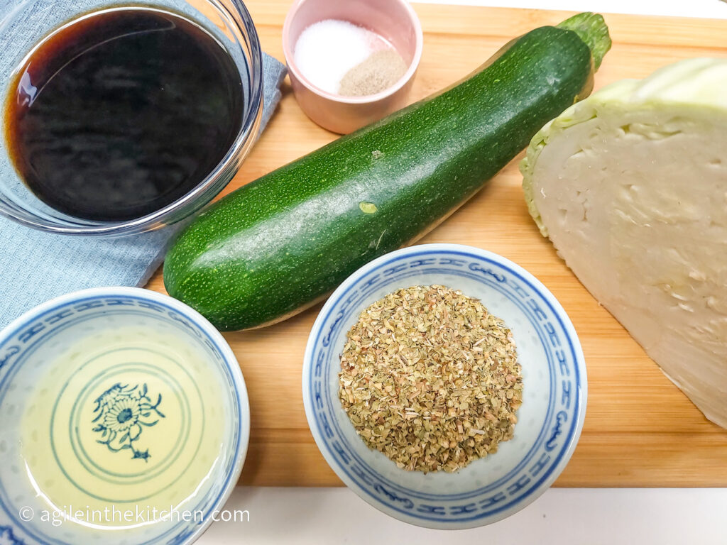 On a wooden cutting board ingredients to make cabbage salad. Clockwise form top left corner: A glass bowl of balsamic vinegar, a small pink bowl with salt and white pepper, one whole zucchini, a hlaf head of cabbage, a bowl of oregano, a bowl of white vinegar.