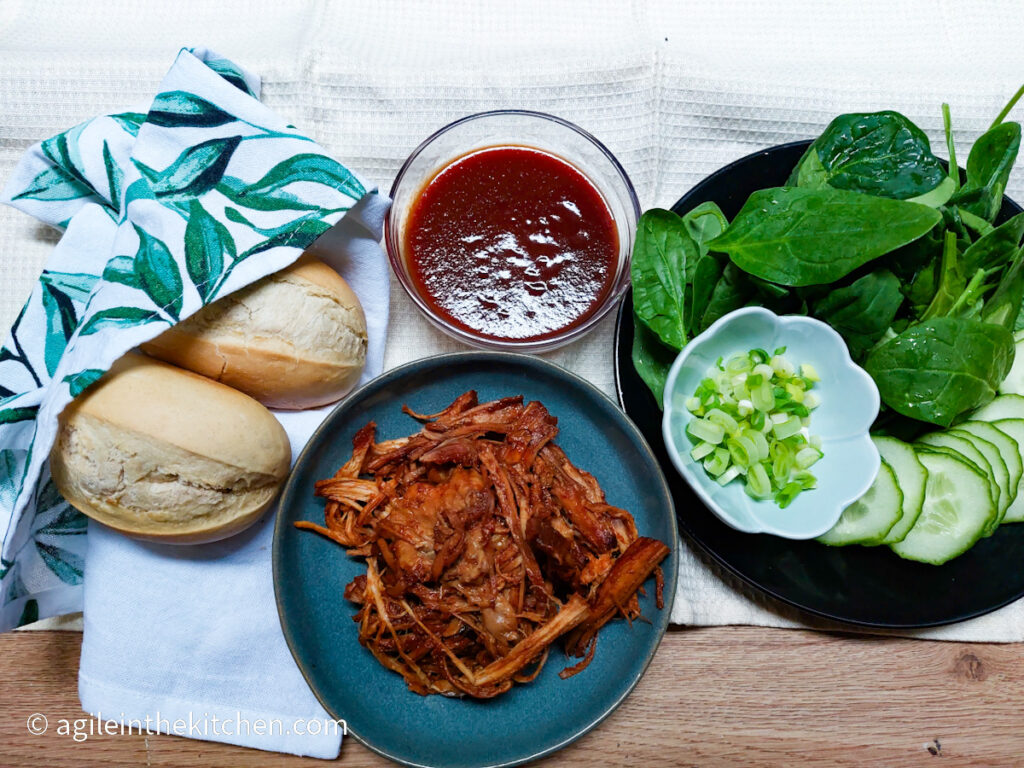 Ingredients to make pulled pork sandwiches. Clockwise from left two mini baguettes wrapped in a kitchen towel, barbecue sauce, a black plate with washed spinach leaves, sliced cucumber and cu green onions, a blue plate piled with pulled pork.