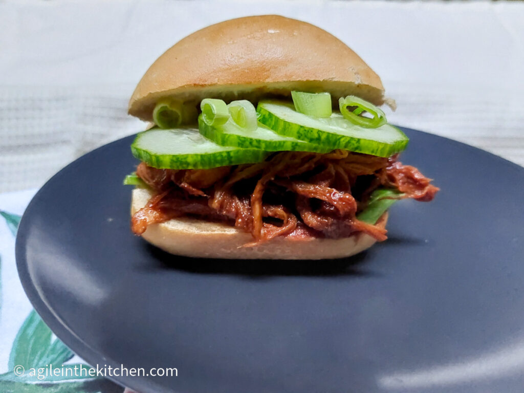 A matte grey plate with a pulled pork sandwich baguette with pulled pork on the bottom, then sliced cucumber, topped with green onions.