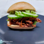 A matte grey plate with a pulled pork sandwich baguette with pulled pork on the bottom, then sliced cucumber, topped with green onions.