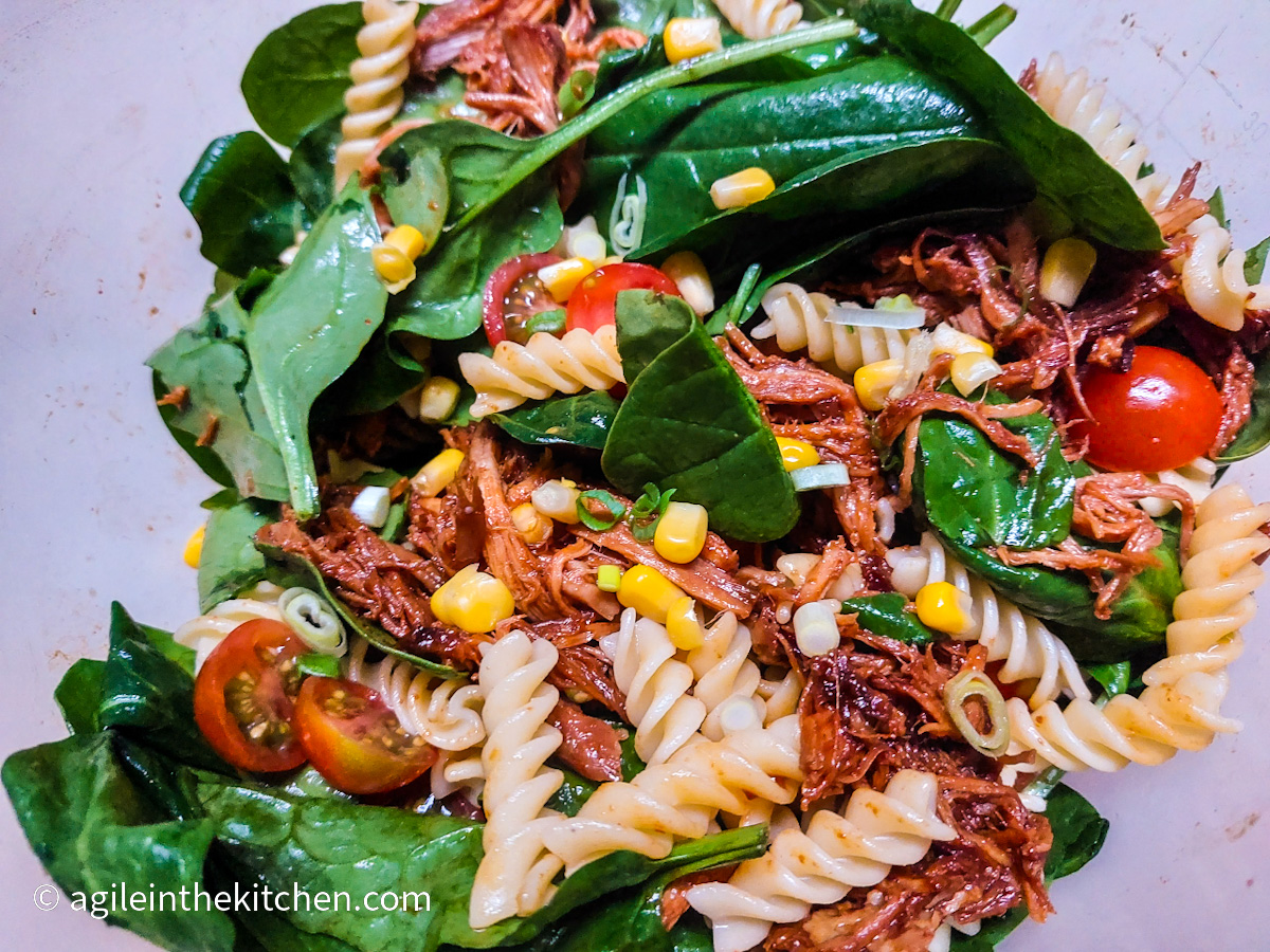 In a bowl, a close up of pulled pork pasta salad with pasta, canned corn, half cut tomatoes, spinach leaves and pulled pork
