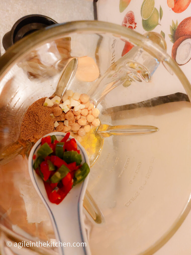 A photo taken from above, looking down into a standing mixer where the ingredients to make hummus is added, a ceramic spoon with chopped chili peppers is about to be added to the other ingredients.