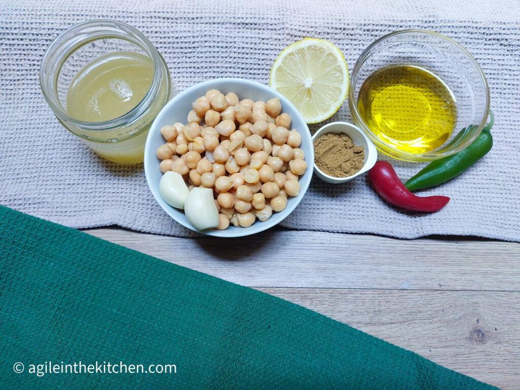 A white textured background with a green textured table cloth in the left hand corner. Ingredients are laid out from the left, a jar of faba water, a bowl of chickpeas and two garlic cloves, half a slice of lemon, a small pot of cumin, a glass bowl of olive oil, a red chili pepper and a green chili pepper.