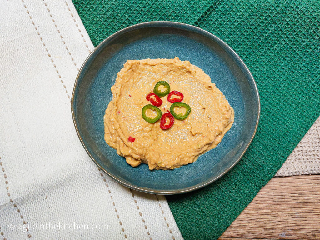 On a green textured background, with a linen table cloth in the left hand corners, a blue plate in the middle with spicy hummus smeared on the plate, topped with a few sliced chilipeppers