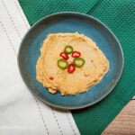 On a green textured background, with a linen table cloth in the left hand corners, a blue plate in the middle with spicy hummus smeared on the plate, topped with a few sliced chilipeppers