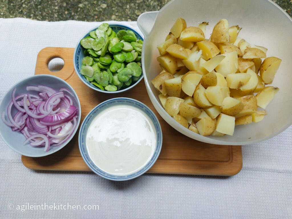 A wooden cutting board on a white background, ingredients to make potato salad laid out. Clockwise from left; a bowl of sliced red onions, a bowl of green broad beans, a large bowl of cooked and cut potatoes, a bowl of mixed potato salad dressing.