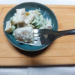 A wooden cutting board on a white background, a blue plate with potato salad, in the foreground a fork with a piece of potato on it