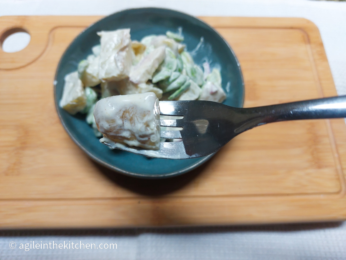 A wooden cutting board on a white background, a blue plate with potato salad, in the foreground a fork with a piece of potato on it
