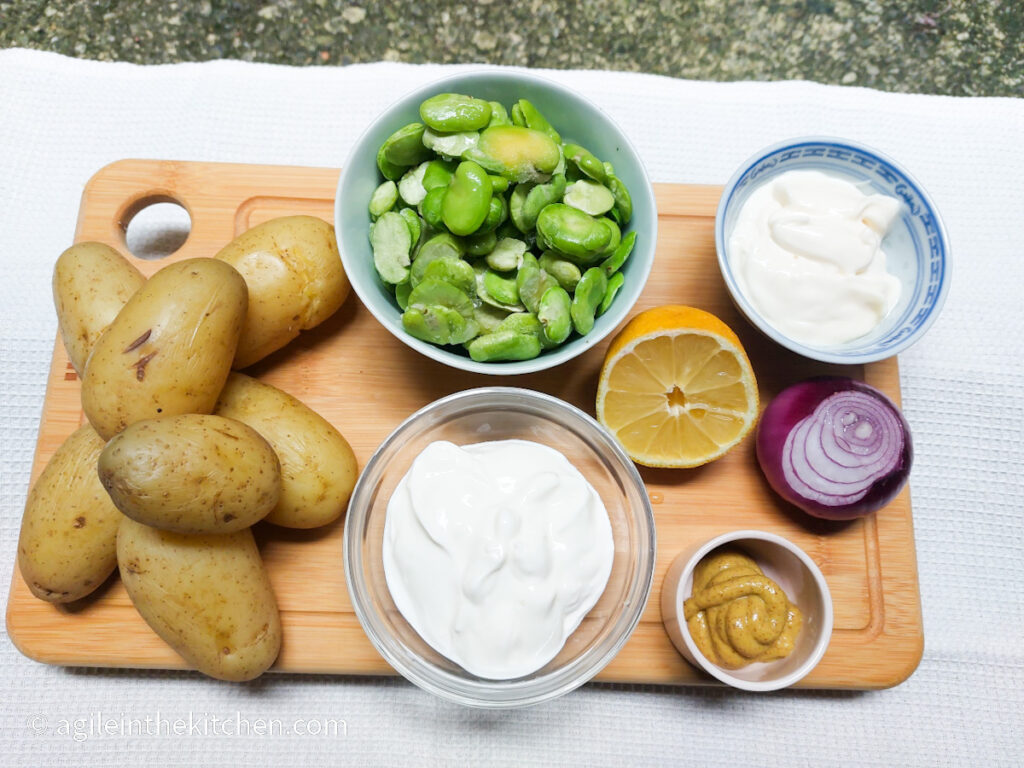 A wooden cutting board on a white background, ingredients to make potato salad laid out. Clockwise from left; cooked potatoes, a bowl of green broad beans, cut lemon, a bowl of mayonnaise, a red onion, a small pot of Dijon mustard, a bowl of Greek yogurt.