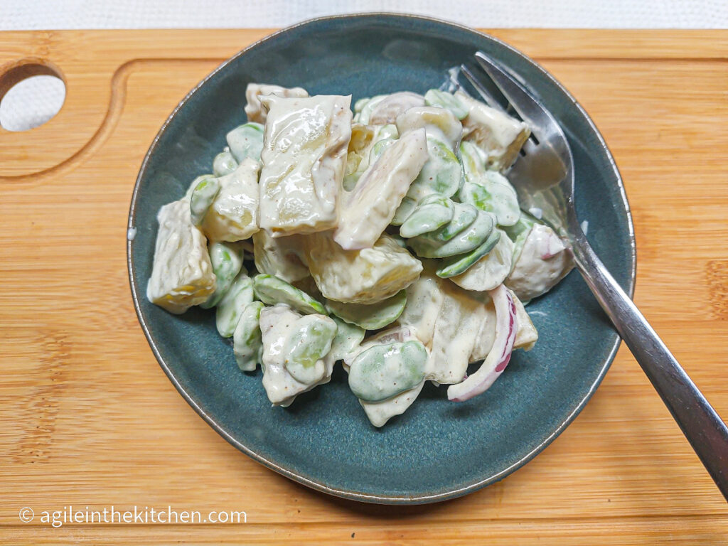 A wooden cutting board on a white background, a blue plate with potato salad and fork on the right hand side.