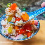 On a wooden cutting board a blue patterned bowl with rainbow risotto, a hand is holding a ceramic spoon with a large pile of risotto towards the centre of the photo.