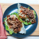 On a wooden cutting board, with a pink cloth napkin on the left side, a blue matted plate with two large salad leaves filled with shredded carrots, white cabbage, Teriyaki tofu, rice noodles and roasted onions.