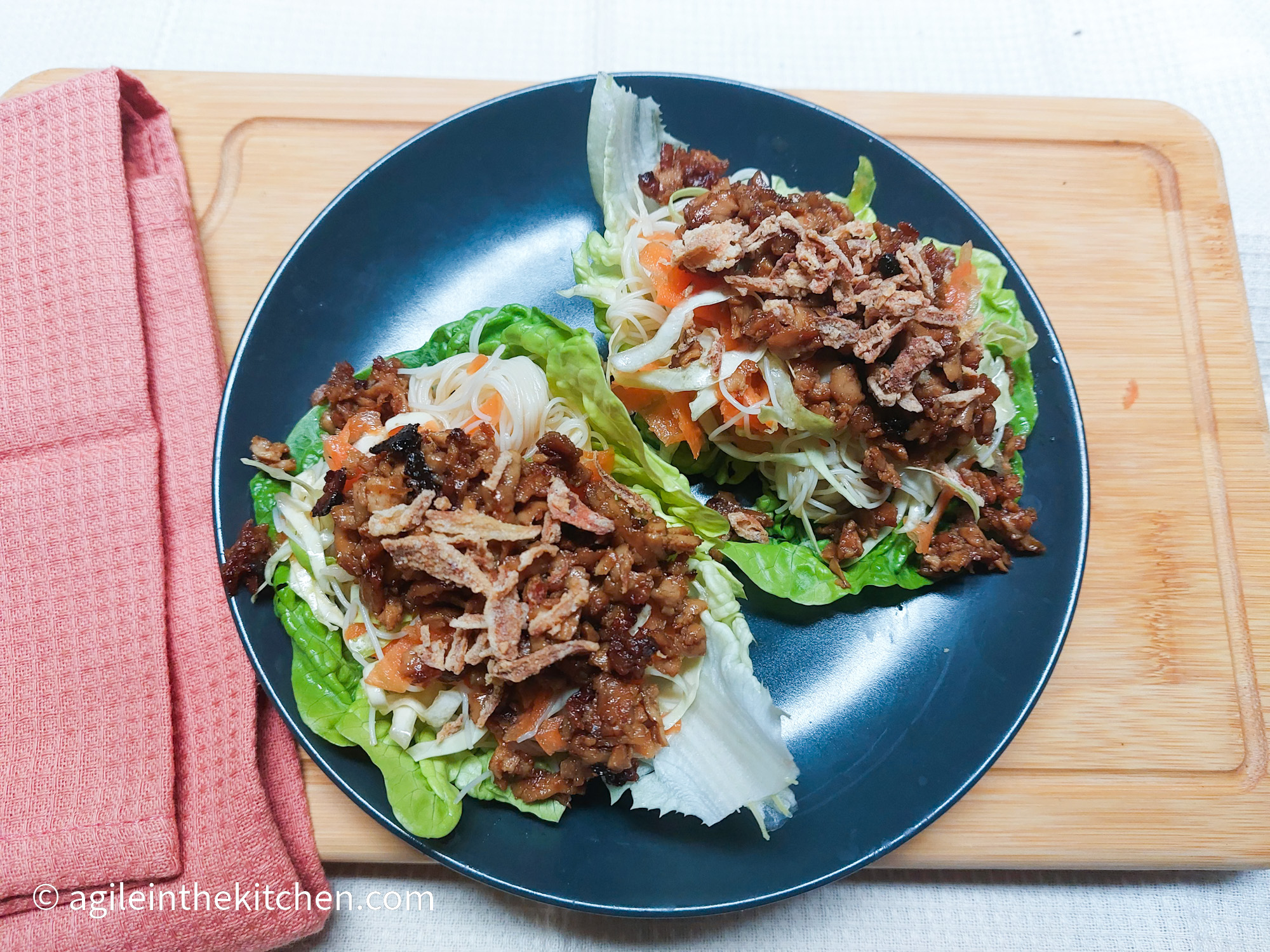 On a wooden cutting board, with a pink cloth napkin on the left side, a blue matted plate with two large salad leaves filled with shredded carrots, white cabbage, Teriyaki tofu, rice noodles and roasted onions.