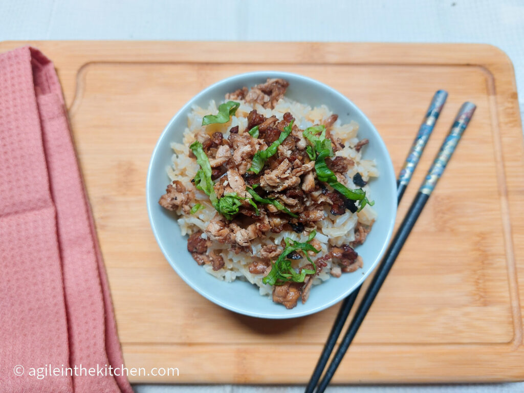 On a wooden cutting board, with a pink cloth napkin on the left side, a bowl of rice topped with shredded bulgogi tofu and a sprinkle of green onions, next to the bowl a pair of chopsticks.