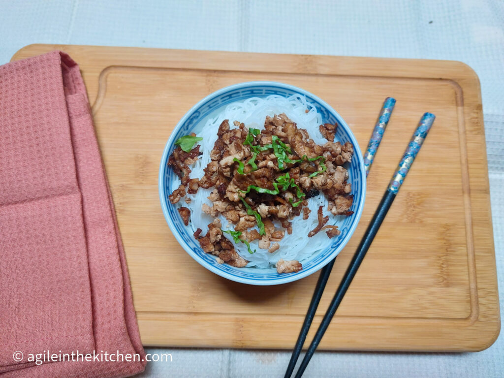 On a wooden cutting board, with a pink cloth napkin on the left side, a bowl of rice noodles topped with shredded bulgogi tofu and a sprinkle of green onions, next to the bowl a pair of chopsticks.