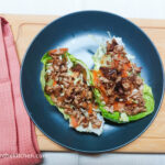 On a wooden cutting board, with a pink cloth napkin on the left side, a blue matted plate with two large salad leaves filled with shredded carrots, white cabbage and shredded bulgogi tofu.