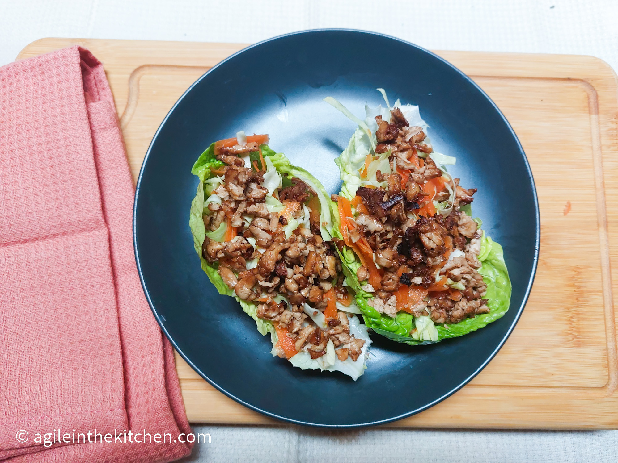 On a wooden cutting board, with a pink cloth napkin on the left side, a blue matted plate with two large salad leaves filled with shredded carrots, white cabbage and shredded bulgogi tofu.