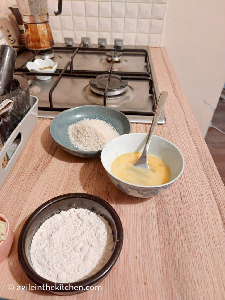 The view from a kitchen, a set up how to make Babybel cheese bites. Closest to the camera a bowl of flour, then a bowl of egg wash, and finally a plate of panko bread crumbs.