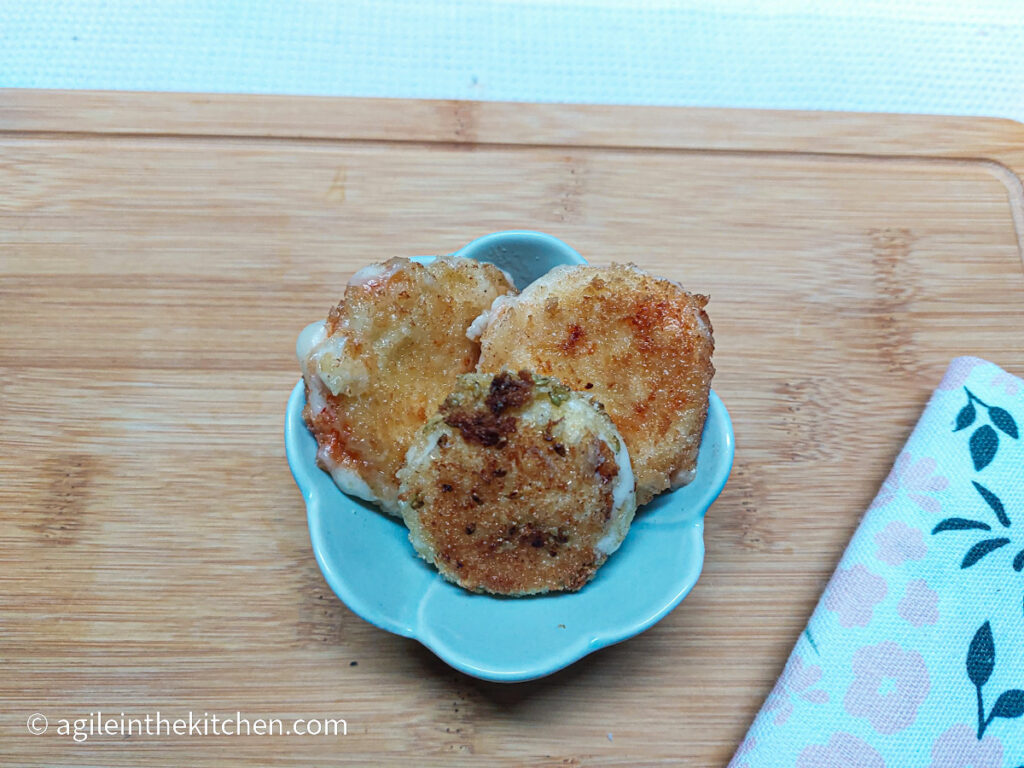 On a wooden cutting board, with a flower patterend cloth napkin in the right corner, a bowl of three panko crusted Babybel cheese bites.