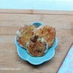On a wooden cutting board, with a flower patterend cloth napkin in the right corner, a bowl of three panko crusted Babybel cheese bites.