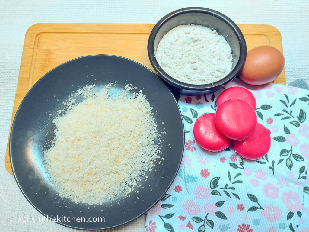 On a white background, a wooden cutting board with a flower patterned cloth napkin in the right lower corner. Clockwise from left, the ingredients to make panko crusted Babybel bites, a plate with panko bread crumbs, a bowl of flour, an egg and four roundels of Babybel cheese,