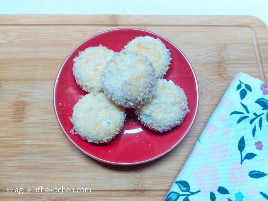 On a wooden cutting board, with a flower patterned cloth napkin in the right corner, a red plate of four panko crusted Babybel cheese bites ready for the freezer.
