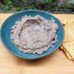 On a wooden cutting board, a photo taken from above, a blue plate with black bean hummus in the middle, to the right hand side a handful of tortilla chips.