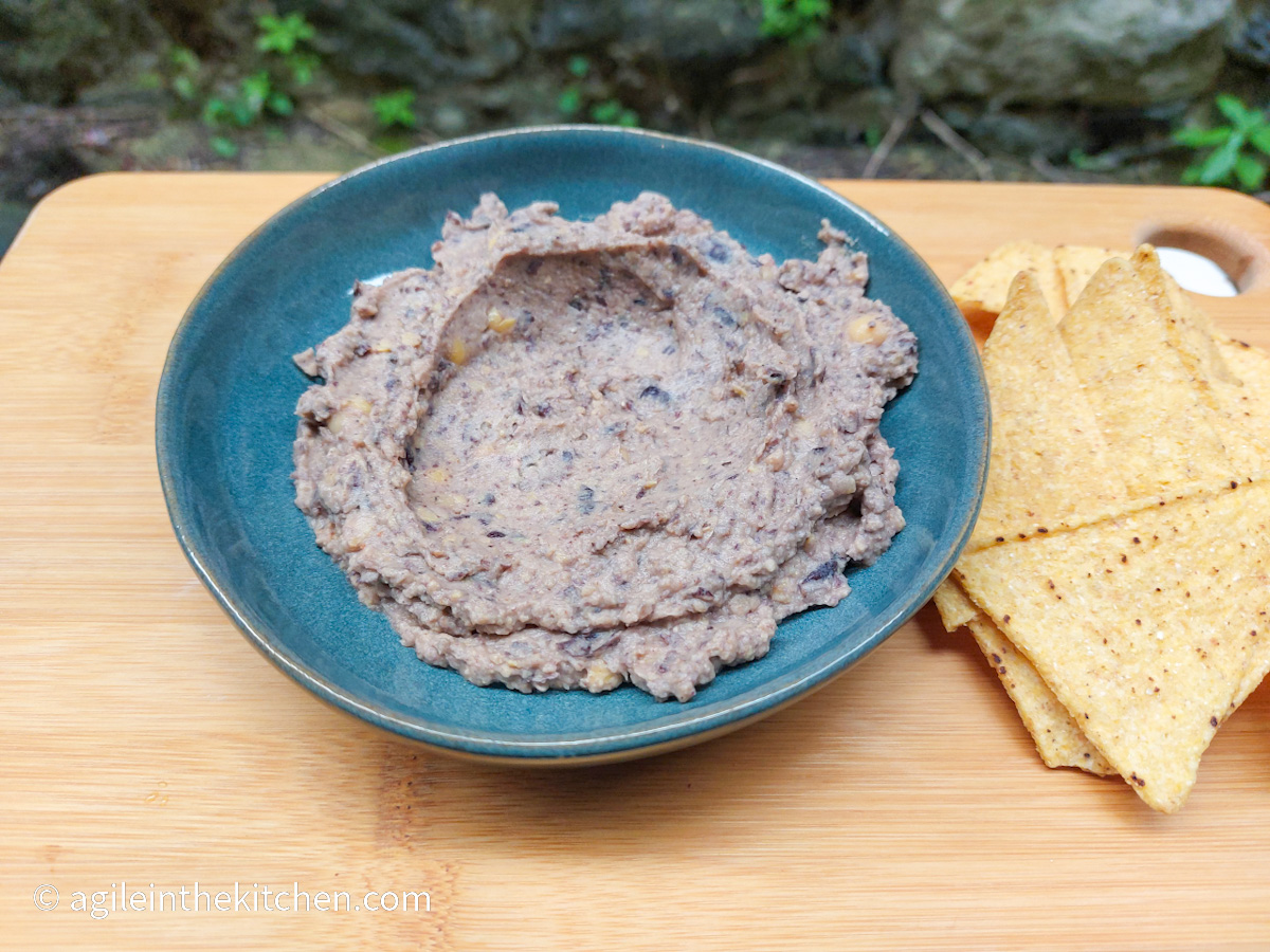 On a wooden cutting board, a photo taken from above, a blue plate with black bean hummus in the middle, to the right hand side a handful of tortilla chips.