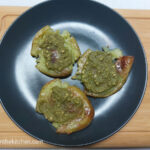 On a white background, a wooden cutting board with a black matte plate with three smashed potatoes with pesto.