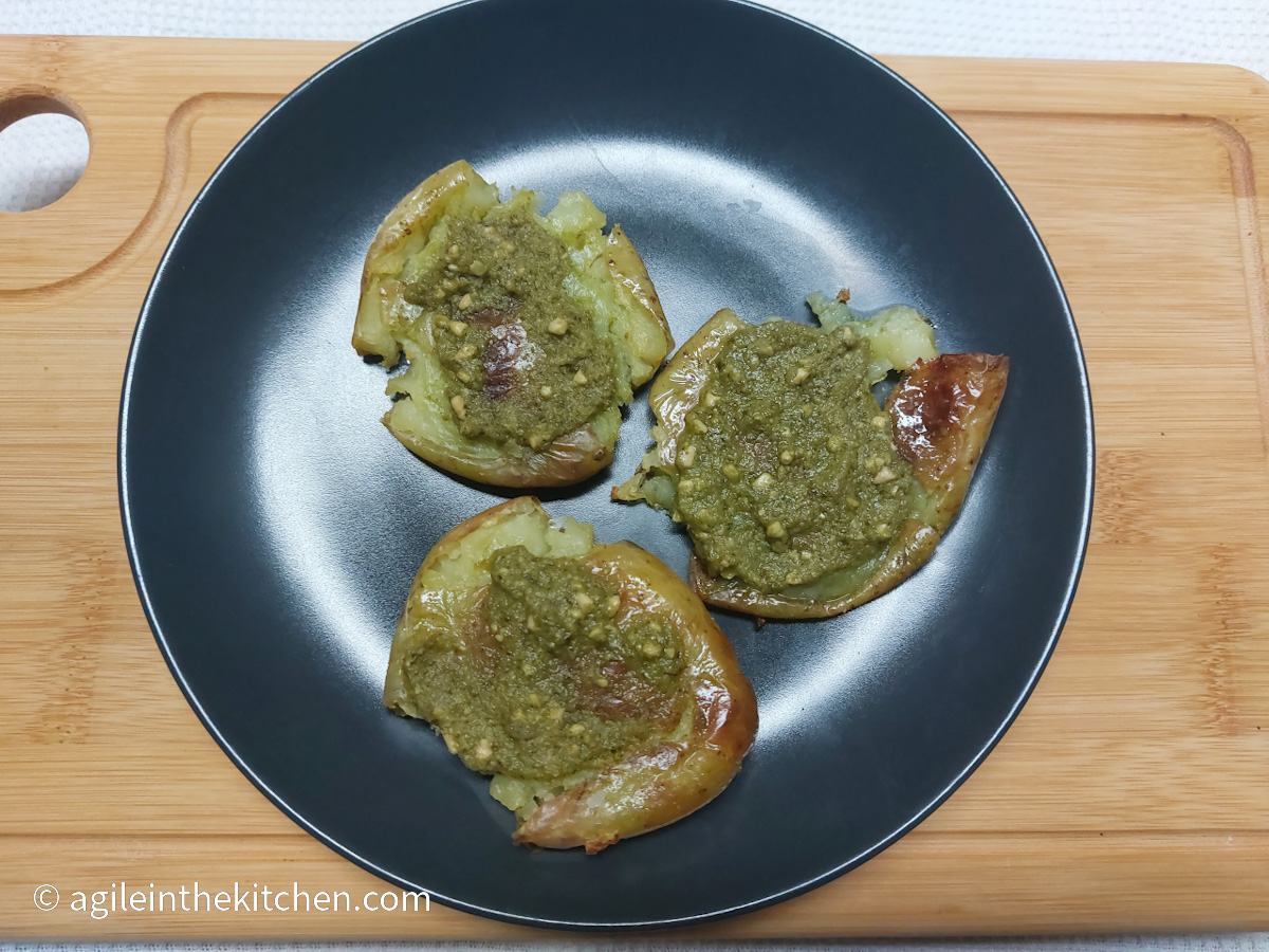 On a white background, a wooden cutting board with a black matte plate with three smashed potatoes with pesto.
