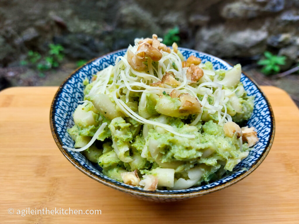 On a wooden cutting board with a blue patterned bowl filled with broccoli mac and cheese, topped with walnuts and shredded cheese.