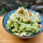 On a wooden cutting board with a blue patterned bowl filled with broccoli mac and cheese, topped with walnuts and shredded cheese.