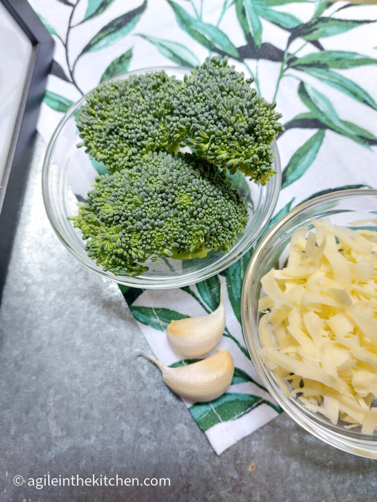 On a silver metallic background, ingredients to make broccoli mac and cheese. Clockwise from left, a bowl of broccoli heads, a bowl of shredded cheese and two garlic cloves.