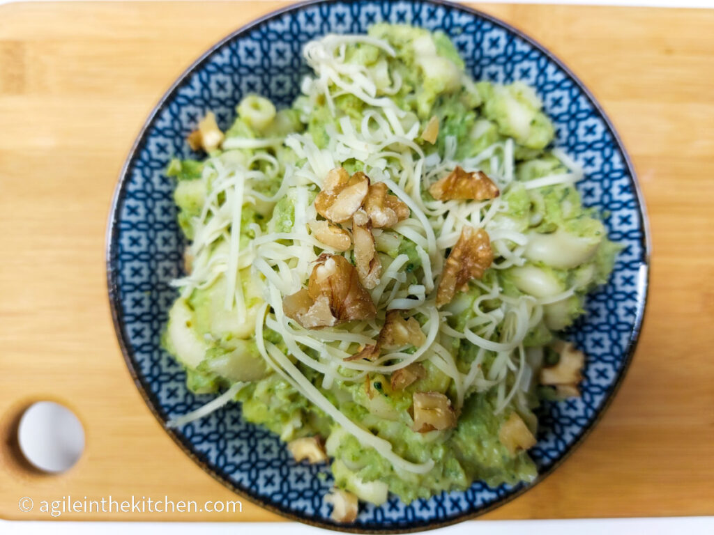 A photo taken from above on a wooden cutting board with a blue patterned bowl filled with broccoli mac and cheese, topped with walnuts and shredded cheese.