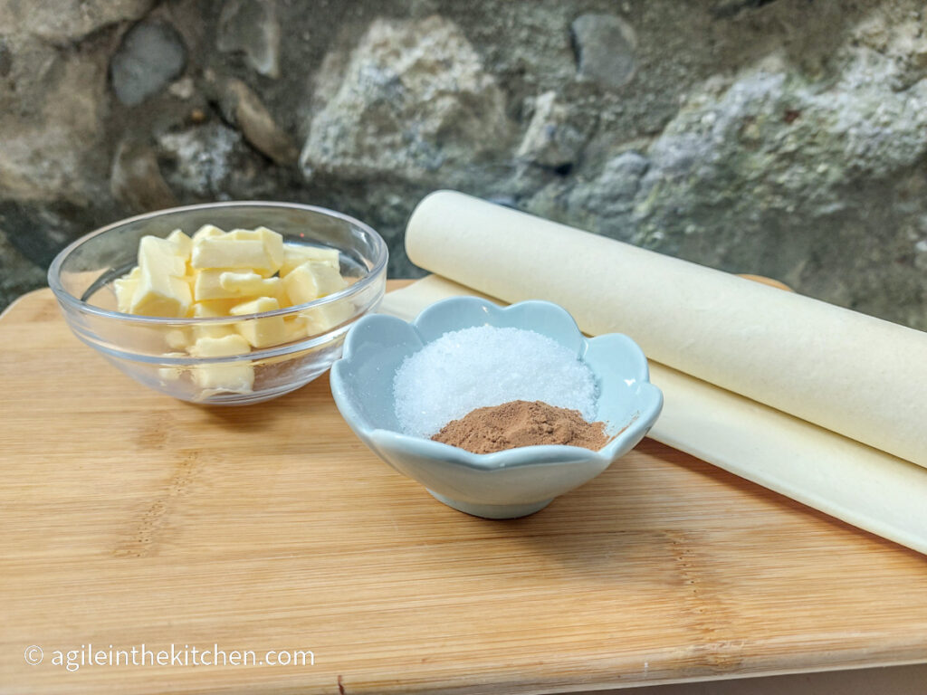 On a wooden cutting board, a glass bowl with cubed butter, a blue smaller bowl with sugar and cinnamon, and a roll of puff pastry.