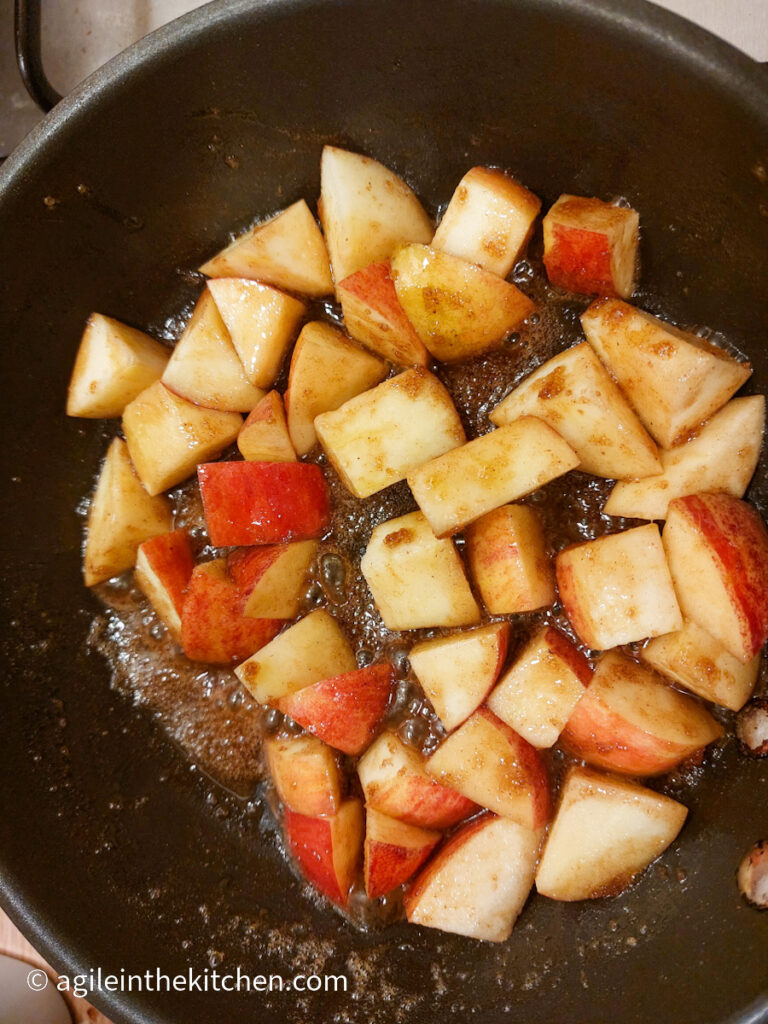 In a frying part, pieces of apples with their red skin still on, cooking in a mixture of butter, cinnamon and sugar