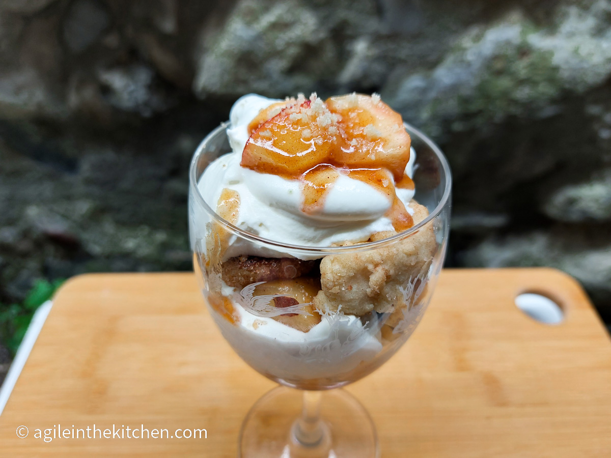 On a wooden cutting board, a tall glass with a layered apple crumble cup with crumble pieces, vanilla cream and apple pieces.