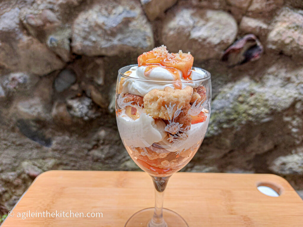 On a wooden cutting board, a tall glass with a layered apple crumble cup with crumble pieces, vanilla cream and apple pieces.