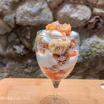 On a wooden cutting board, a tall glass with a layered apple crumble cup with crumble pieces, vanilla cream and apple pieces.